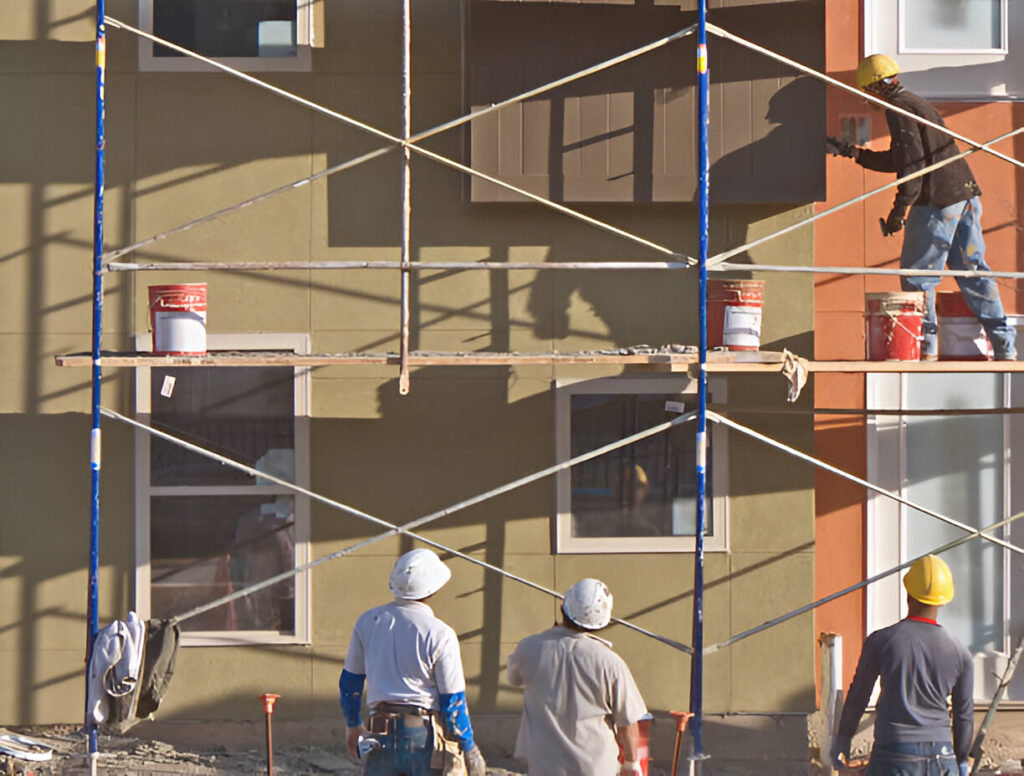 Three supervisors or co-workers watch as a workman on scaffolding does the actual work.