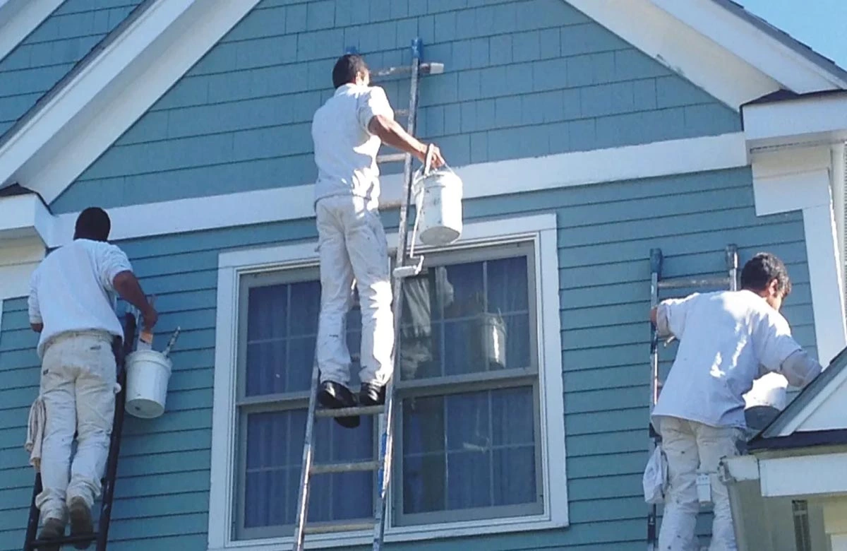 Team of painters using ladders to paint the upper level of a blue wooden house.