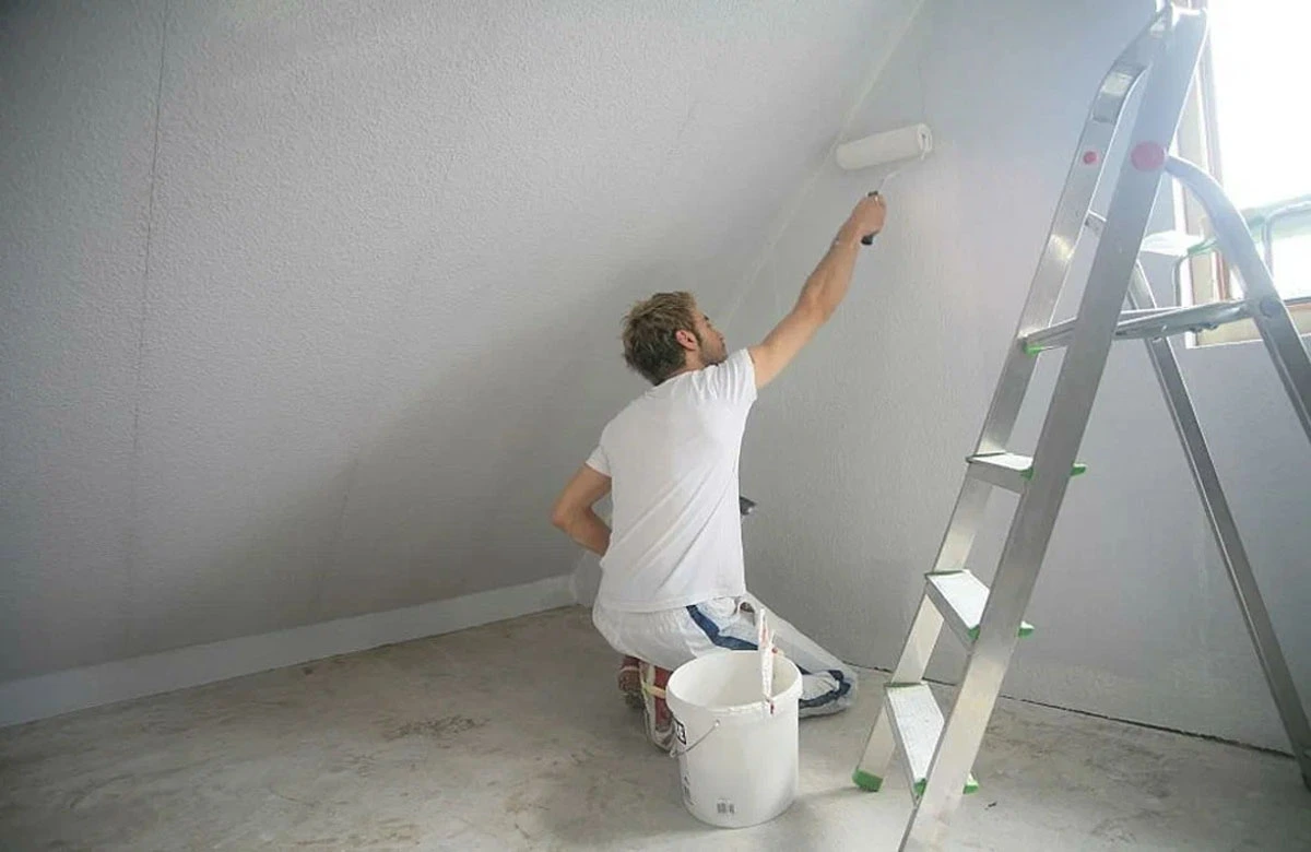 A painter in a white shirt uses a roller to paint the ceiling while kneeling next to a ladder and paint bucket, reflecting the skill and dedication of professional home painters in Dubai.
