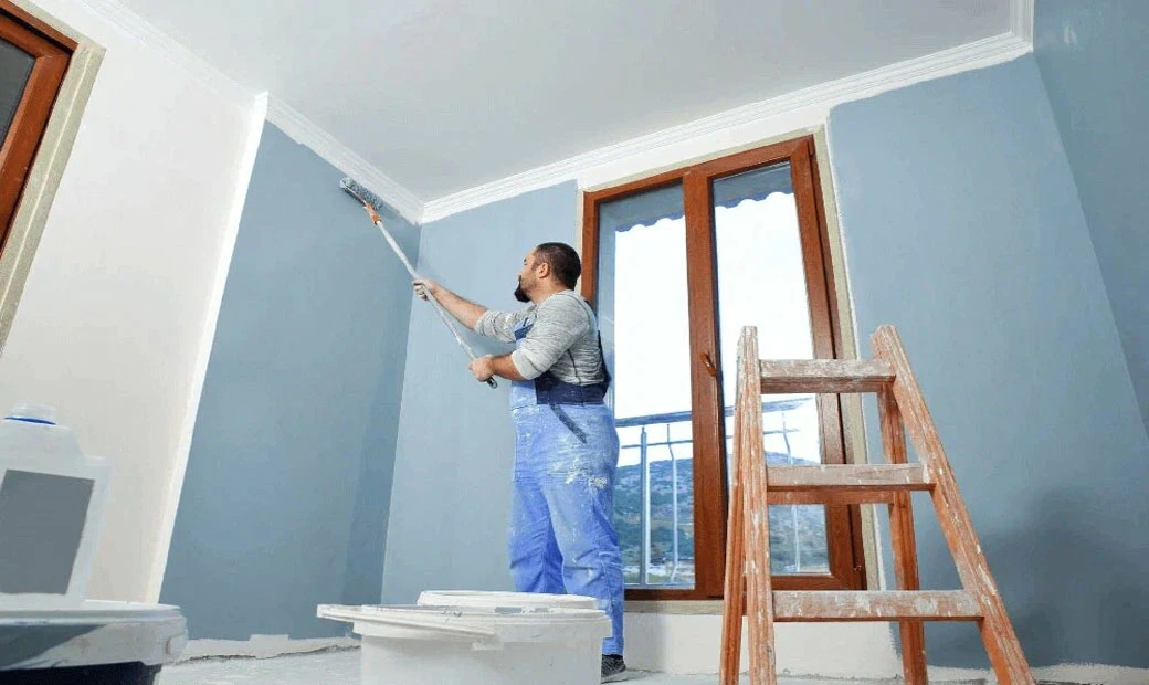 Painter in blue overalls painting a wall blue inside a modern room with large windows.