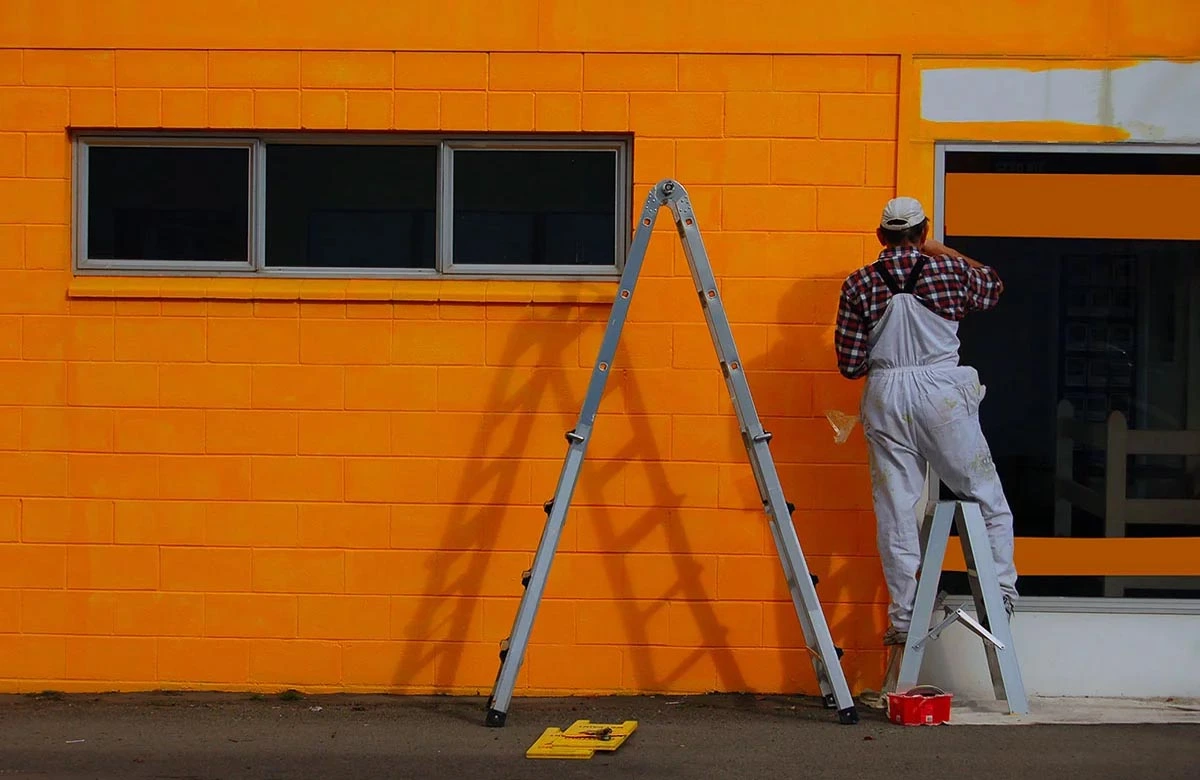 Painter applying bright orange paint on a commercial building exterior with a roller.