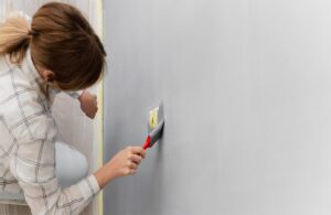 Woman carefully painting around a socket on a grey wall using a brush — wall paint Dubai.