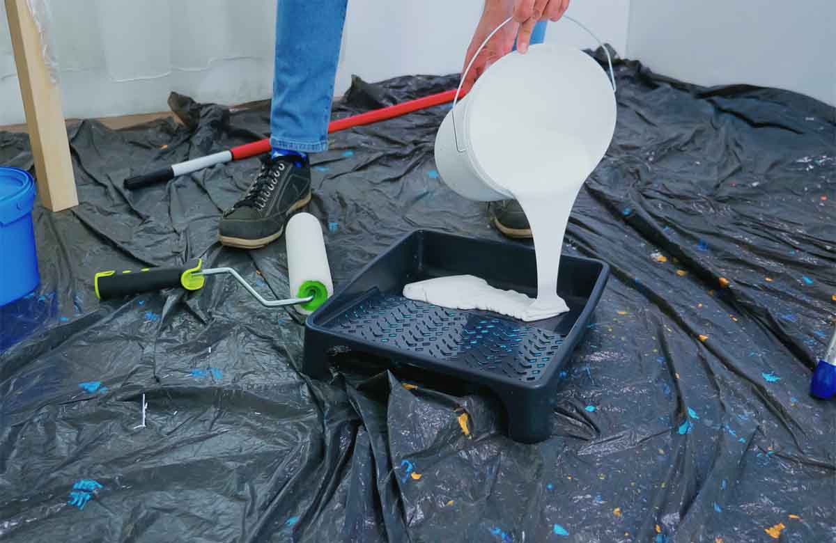 Worker pouring white paint into a tray on a tarp-covered floor with tools — hire a paint contractor Dubai.