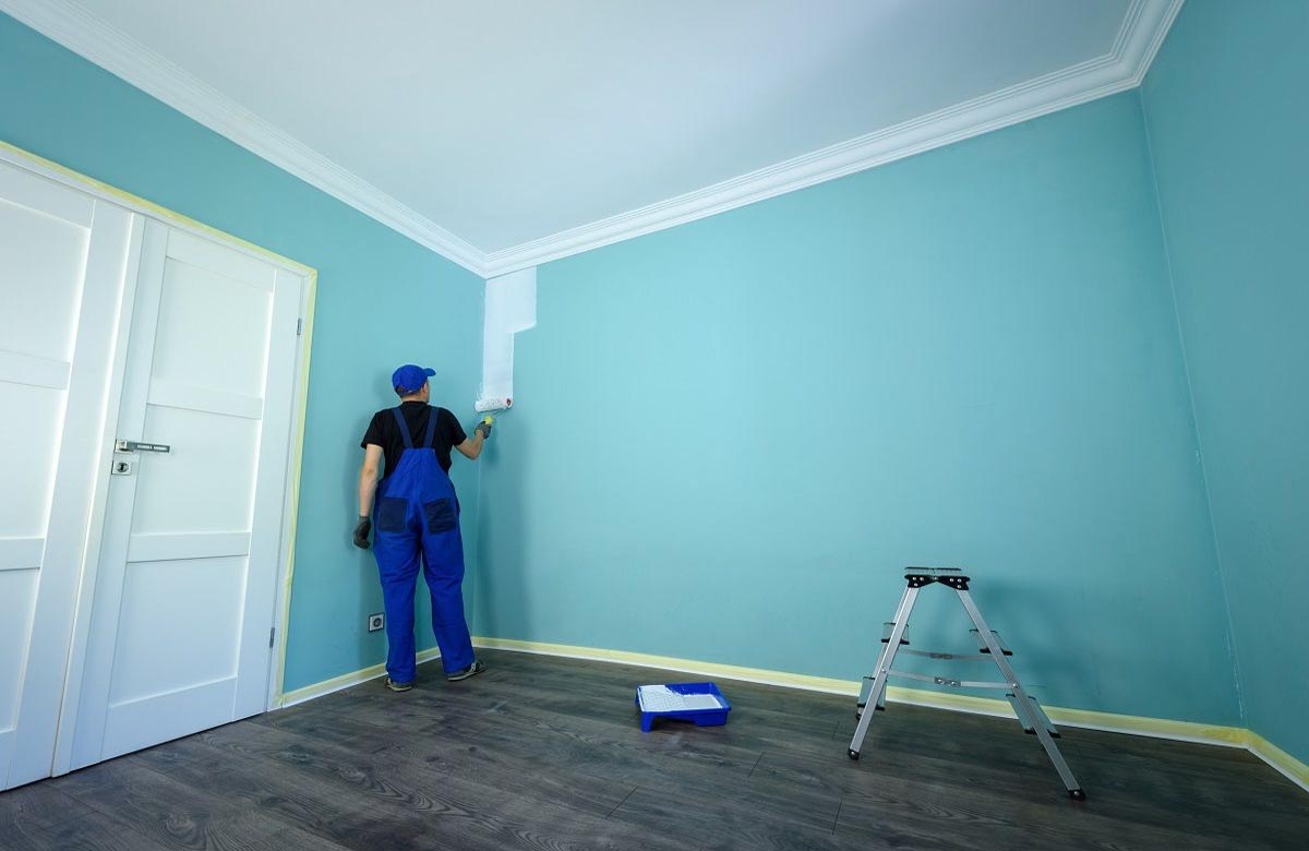 paint finish for Dubai walls | Professional painter in blue overalls painting a teal wall white — quality service by top Interior Paint Companies in Dubai.