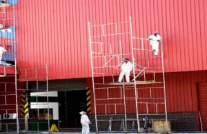 Workers providing industrial painting services in Dubai, painting a large red metal building using scaffolding.