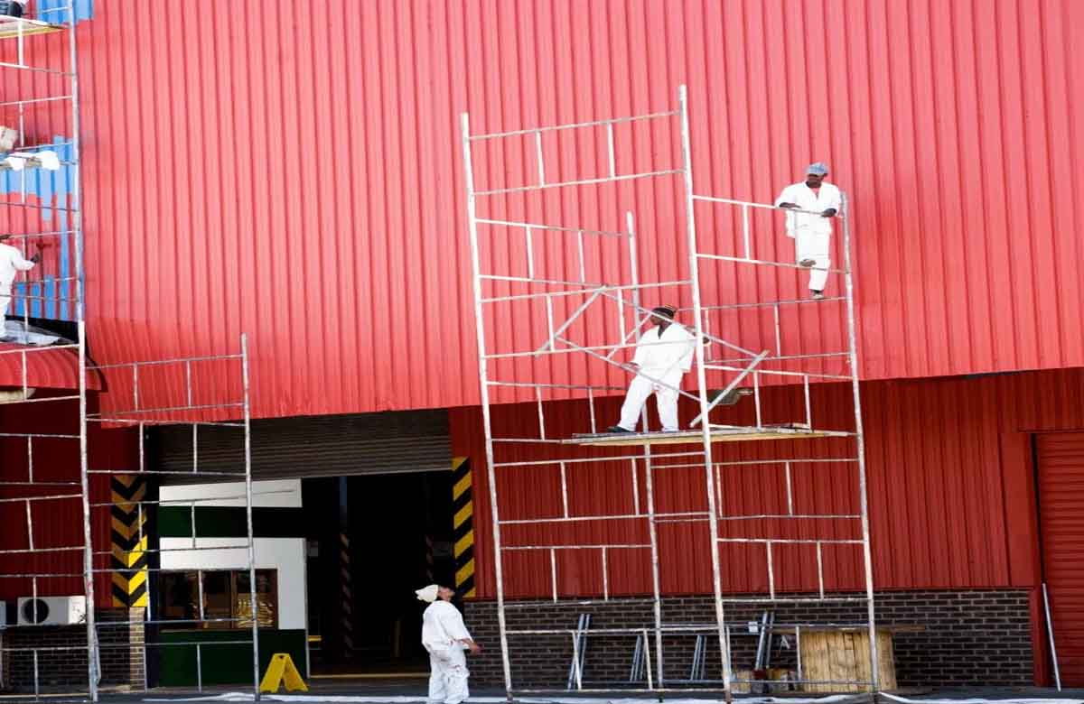 Workers providing industrial painting services in Dubai, painting a large red metal building using scaffolding.