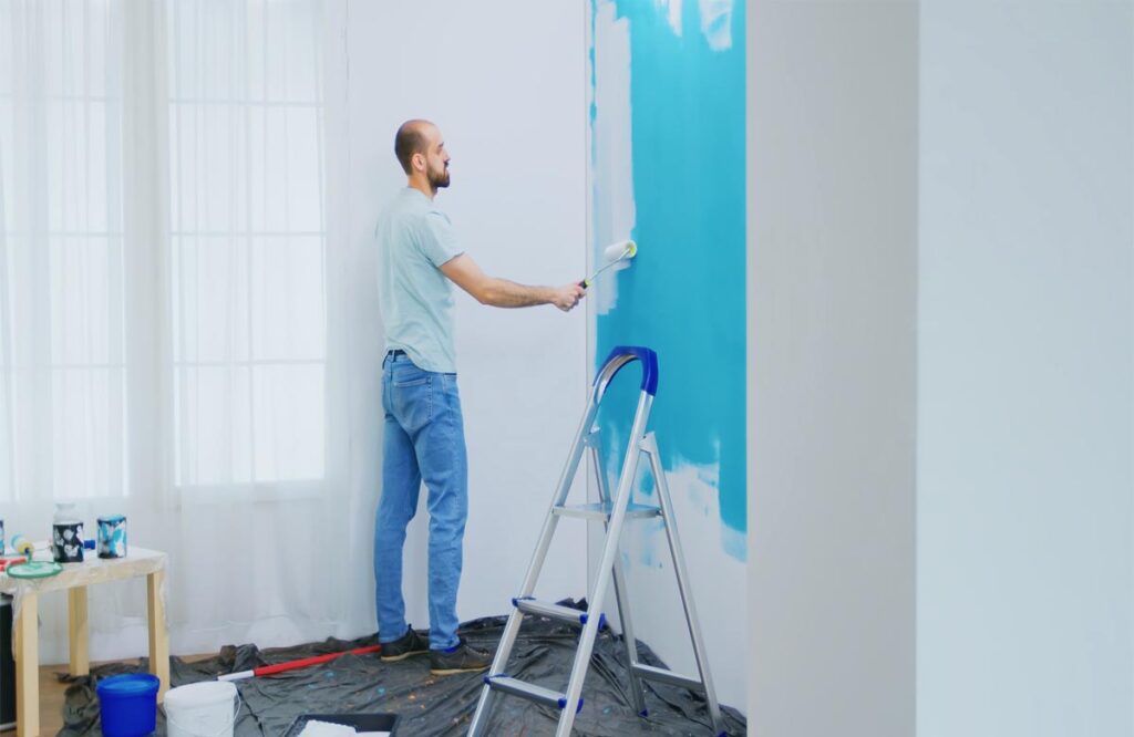 Man painting a white wall with blue color using a roller brush near a ladder — Interior Paint Company in Dubai.