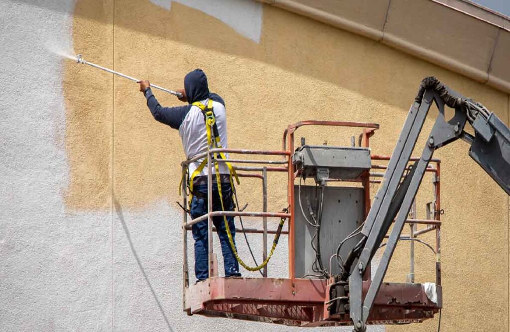 Industrial Painting Contractors in Dubai spraying white paint on a tall yellow building using a boom lift for safe and efficient exterior work.